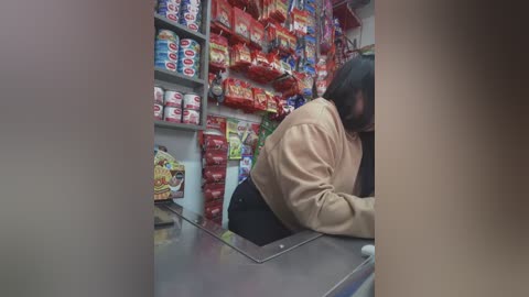 Media: A blurry video of a woman with long black hair, wearing a beige jacket, leaning over a checkout counter in a brightly lit convenience store. The background is filled with shelves stocked with colorful snacks and beverages.