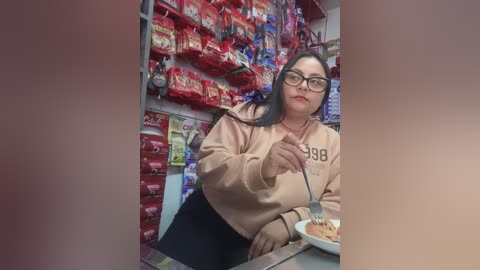 Video of a middle-aged woman with long dark hair, glasses, and a beige sweatshirt, eating at a supermarket counter. Background shows red and yellow packaged foods.