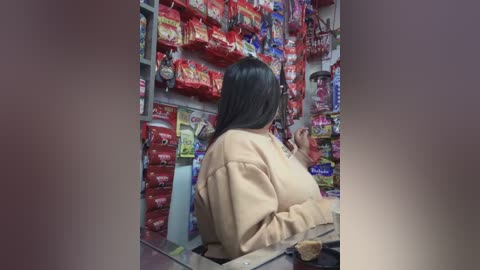 Media: A video of a cashier with long, straight black hair, wearing a beige sweater, standing behind a glass counter, surrounded by colorful, stacked snack packages in a bustling store.