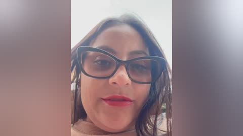 Media: Video of a woman with light brown skin and shoulder-length dark hair, wearing black-rimmed glasses and red lipstick, set against a blurred background, creating a soft, dreamy effect.