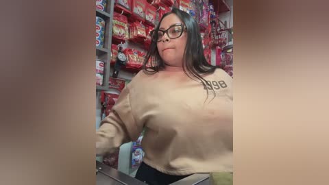 Media: Video of a plus-sized woman with long dark hair, wearing glasses and a beige sweatshirt, shopping in a brightly lit grocery store aisle filled with colorful snacks.