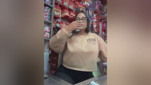 Media: Video of a plus-sized woman with glasses, wearing a beige sweatshirt with \"1988\" and black leggings, taking a selfie in a colorful, crowded convenience store aisle filled with snacks.