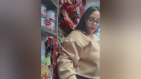 A video of a woman with glasses, wearing a beige sweater, standing in a grocery store aisle with red and white bags of chips and a shelf of white containers.