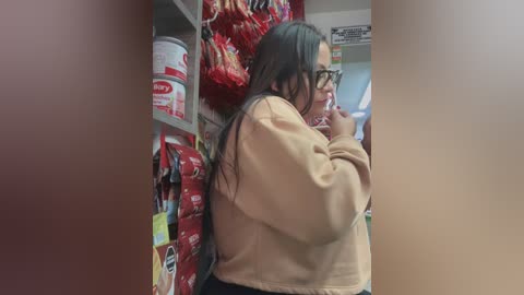 Video of a woman in a beige jacket, with long dark hair, glasses, and a contemplative expression, standing in a grocery aisle filled with red and white packaged goods.