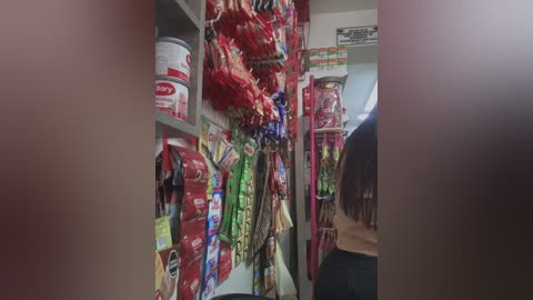 A video of a grocery store aisle with a woman, back to the camera, reaching for snacks. Brightly colored chips, cookies, and cereals line the shelves, with a visible red and white \"M&M's\" container on the left.