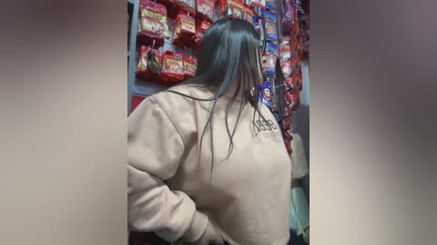 Media: Video of a woman with long, straight black hair, wearing a beige hoodie, shopping in a brightly lit grocery store. She is looking at the shelves filled with various snack items, including red and yellow bags.