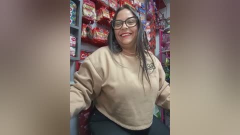 Media: Video of a plus-sized woman with long black hair, glasses, and a beige sweater, smiling in a brightly lit grocery aisle filled with colorful snacks.