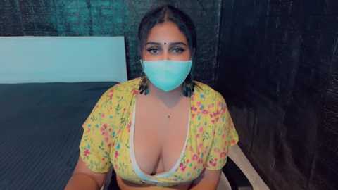 Media: Video of a curvy, South Asian woman with light brown skin, large breasts, wearing a yellow floral blouse, blue face mask, and braid, sitting on a bed with dark walls in the background.