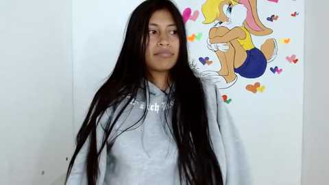 Media: Video of a young woman with long, black hair in a gray hoodie, standing against a white wall adorned with a colorful, cartoonish drawing of a bunny and hearts.