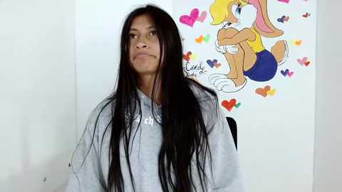 Media: Video of a young woman with long black hair, wearing a gray hoodie, standing in front of a colorful, cartoonish background featuring Bugs Bunny and Lola Bunny.