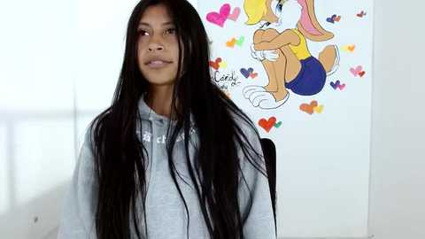 Media: Video of a young Black woman with long black hair, wearing a grey hoodie, sitting indoors. Background features a colorful, cartoonish drawing of Lola Bunny on a white wall.