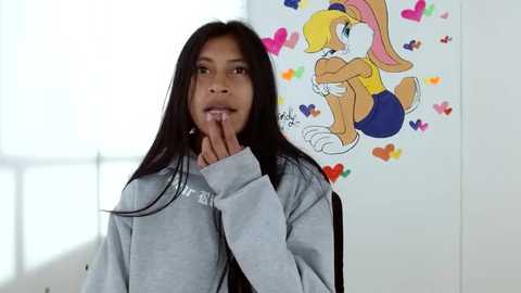 A young girl with long black hair, in a gray sweatshirt, stands in a brightly lit room with a colorful Looney Tunes-themed wall mural, touching her lips.