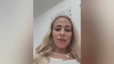 Media: A video of a blonde woman with long, wavy hair, wearing a white top and pearl necklace, standing against a white wall with a framed document.