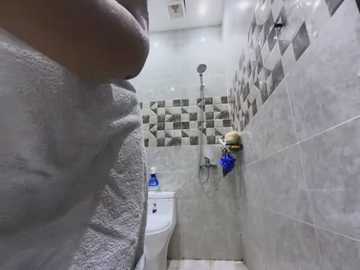 Media: Video of a narrow, modern bathroom with white and gray tiled walls, a white toilet on the left, and a person with a white towel wrapped around their waist.