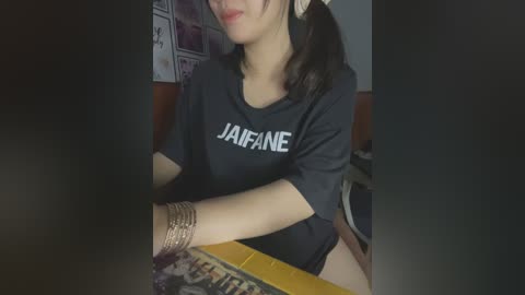 Media: Video of a young Asian woman with light skin, wearing a black \"JAFANE\" t-shirt, seated at a table with a yellow surface. She has long black hair and wears multiple bracelets on her right wrist. The background includes a wall with posters and a dark, blurry setting.