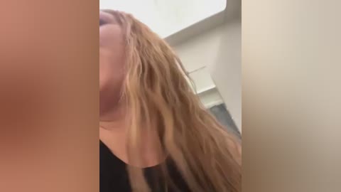 Media: Video of a close-up of a woman with long, wavy blonde hair, seen from the side, partially obscuring her face, in a light-colored room.