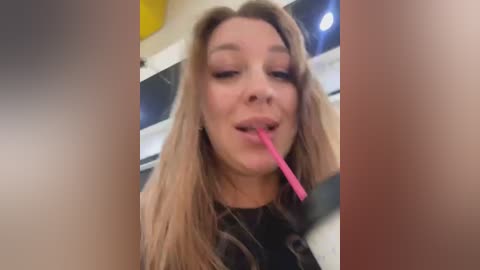 Media: Video of a young woman with light skin, long blonde hair, wearing a black top, licking a pink straw, indoors, blurry background.