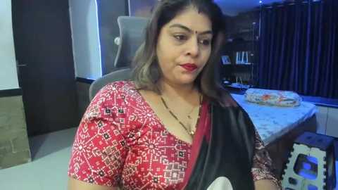 Media: Video of a middle-aged South Asian woman with shoulder-length dark hair, wearing a red and black patterned sari, seated indoors, in a modern, dimly lit room with a white bed and bookshelves.