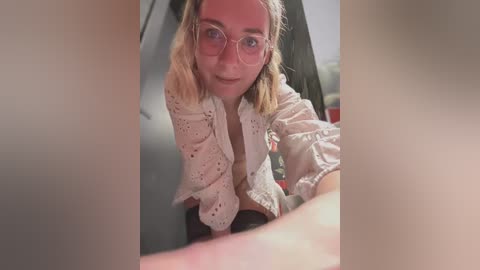 Media: Video of a blonde woman with glasses, wearing a white lace top, kneeling in a dimly lit room, looking directly at the camera.