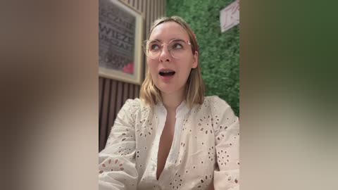 Media: Video of a blonde woman with glasses, wearing a white, eyelet-patterned blouse, and a surprised expression. Background includes a green wall and a framed sign.