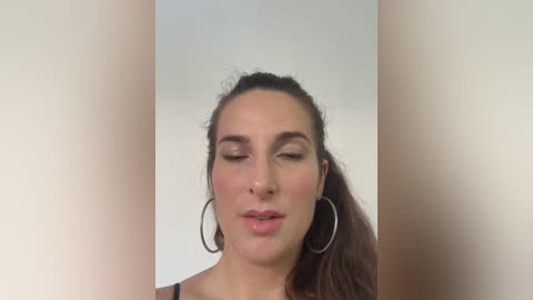 Media: Video of a young woman with fair skin, brown hair in a high ponytail, wearing large hoop earrings, and closed eyes, against a plain beige background.