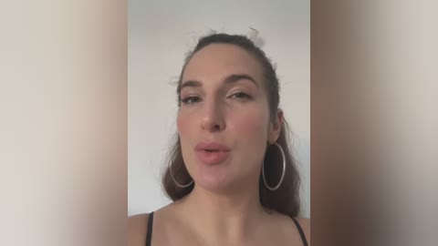 Media: A video of a fair-skinned woman with light brown hair pulled back, wearing a black spaghetti-strap top and large silver hoop earrings. She puckers her lips playfully against a plain, beige background.