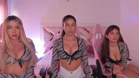 Media: Video of three young women with different skin tones and hair colors, dressed in revealing tops, sitting on a bed with pink pillows in a dimly lit room.