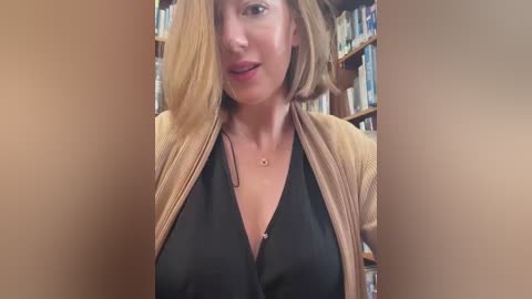 Media: A video of a fair-skinned woman with short, light brown hair, wearing a black blouse and beige cardigan, standing in a library with bookshelves in the background.