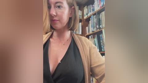 Media: Video of a light-skinned woman with short, straight, light brown hair, wearing a black top and beige cardigan, standing in a library with bookshelves in the background.