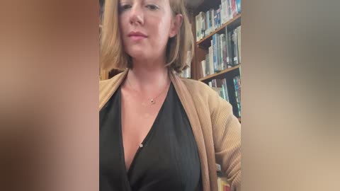 Media: A video of a light-skinned woman with shoulder-length blonde hair, wearing a black top and beige cardigan, standing in a library with bookshelves in the background.