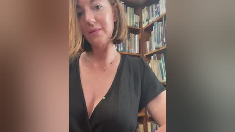 Media: Video of a fair-skinned woman with shoulder-length blonde hair, wearing a black, low-cut blouse, standing in a book-filled room.