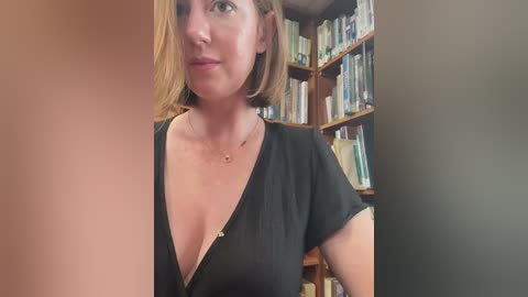 Media: Video of a middle-aged woman with shoulder-length blonde hair, wearing a black V-neck t-shirt, standing in a library with wooden bookshelves filled with books in the background.