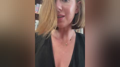 Media: Video of a fair-skinned woman with shoulder-length blonde hair, wearing a black V-neck top, and a delicate necklace. Background shows bookshelves and soft lighting, giving a cozy, indoor ambiance.