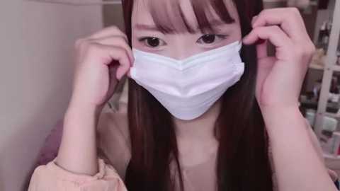 Media: Video of an Asian woman with long brown hair and bangs, wearing a white surgical mask, adjusting it with both hands. She has light skin and is dressed in a light-colored top. The background is blurred, showing shelves with various items.