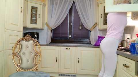 Video of a light beige kitchen with cream cabinets, a decorative chair with a tufted back, and a person in pink leggings and white shirt standing by a window with sheer curtains.