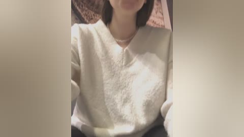 Video of a person with light skin, wearing a white, textured sweater and a pearl necklace, seated in a dimly lit room with a brown rug and beige walls.