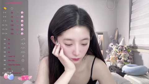 Media: Video of an Asian woman with long black hair, wearing a black top, resting her chin on her hand, in a softly lit room with plush toys and a window.