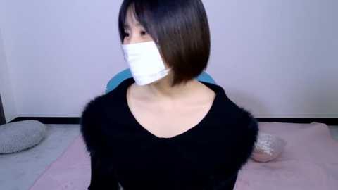 Video of an East Asian woman with straight, shoulder-length black hair, wearing a black sweater with fluffy white fur sleeves, white surgical mask, and sitting on a bed with a pink blanket. The background features a plain white wall and a grey pillow.