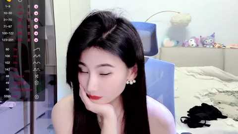 Media: Video of an Asian woman with long black hair, fair skin, and red lipstick, wearing a white top, resting her chin on her hand in a bedroom with a bed, stuffed animals, and a TV.