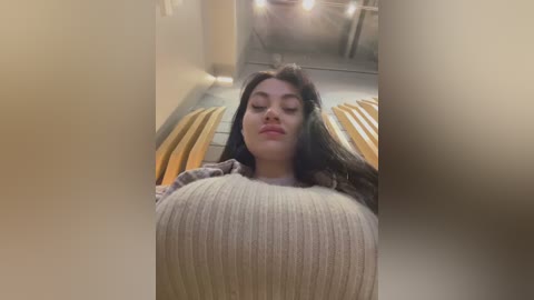 Media: A video of a young woman with long dark hair, wearing a beige ribbed sweater, sitting on an escalator. The background features wooden slats and a ceiling with industrial-style lighting.