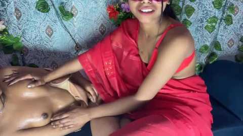 Media: Video of a joyful Indian woman with medium skin tone and a full figure, wearing a red saree, massaging a topless man with a darker skin tone, sitting on a blue bed.