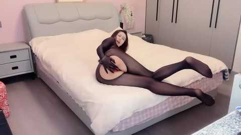 Video of a woman in black tights and a black bodysuit, lying on a bed with light pink walls, revealing her buttocks.