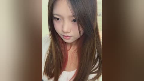 Media: Video of a young Asian girl with straight, shoulder-length brown hair and a pale complexion, wearing a white top. She has a neutral expression and is indoors against a blurred, light-colored background.