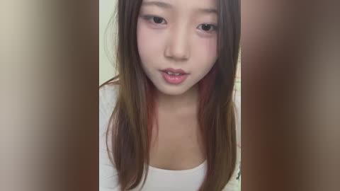 Media: Video of an East Asian woman with long brown hair and red highlights, fair skin, wearing a white top, standing in front of a blurred, neutral-toned background.