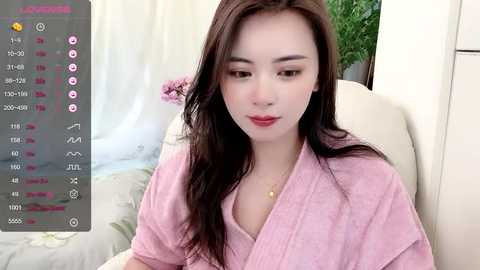 Media: Video of an East Asian woman with long black hair, wearing a pink bathrobe, sitting on a cream couch. The background features a white curtain and a potted plant.