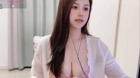 Media: Video of an Asian woman with long brown hair, wearing a sheer white blouse, exposing a pink lace bra, sitting indoors with light-colored curtains in the background.