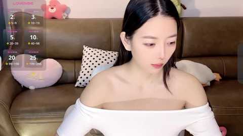 Media: Video of a young East Asian woman with long black hair, wearing a white off-shoulder top, sitting on a brown leather couch. The background features stuffed toys and a calendar.