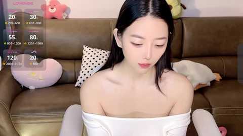 Media: Video of an Asian woman with straight black hair, fair skin, and a slender physique, wearing a white off-shoulder top. She sits on a brown leather couch in a cozy, playful room filled with stuffed toys.