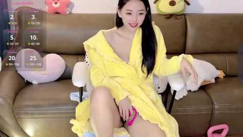 Media: Video of a young Asian woman with long black hair, wearing a yellow robe, sitting on a brown leather sofa with stuffed animals.