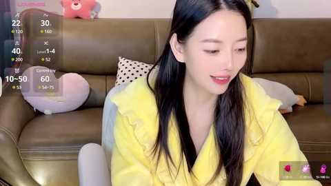 Media: Video of an East Asian woman with long black hair, wearing a yellow coat, sitting on a beige leather couch with stuffed animals in a cozy living room.